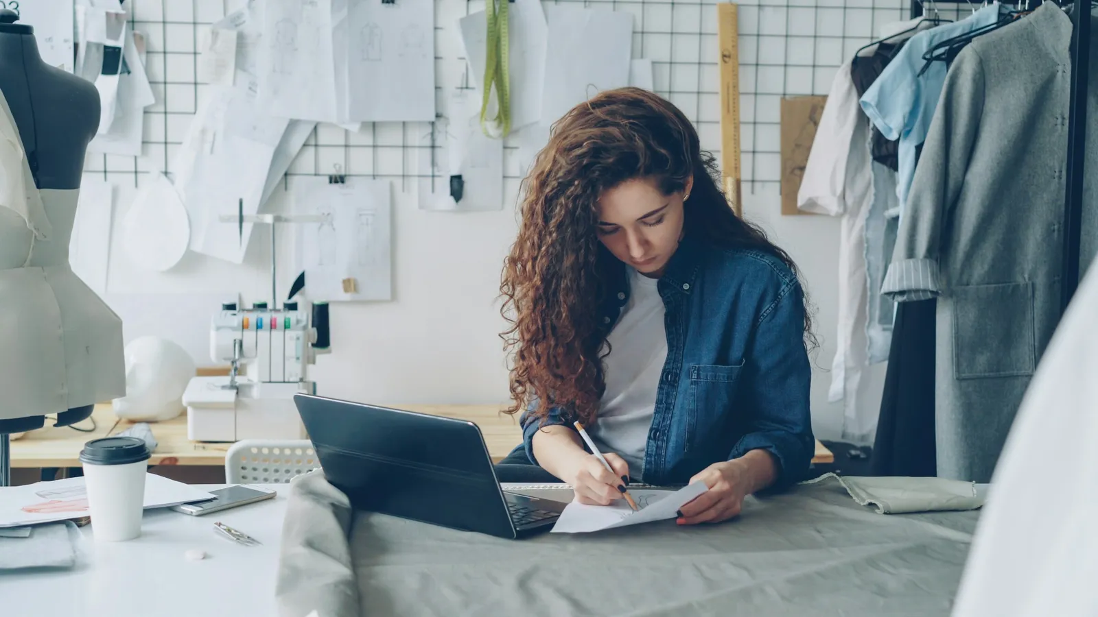 Woman writing on a sheet of paper beside a laptop at a work table in a clothing studio