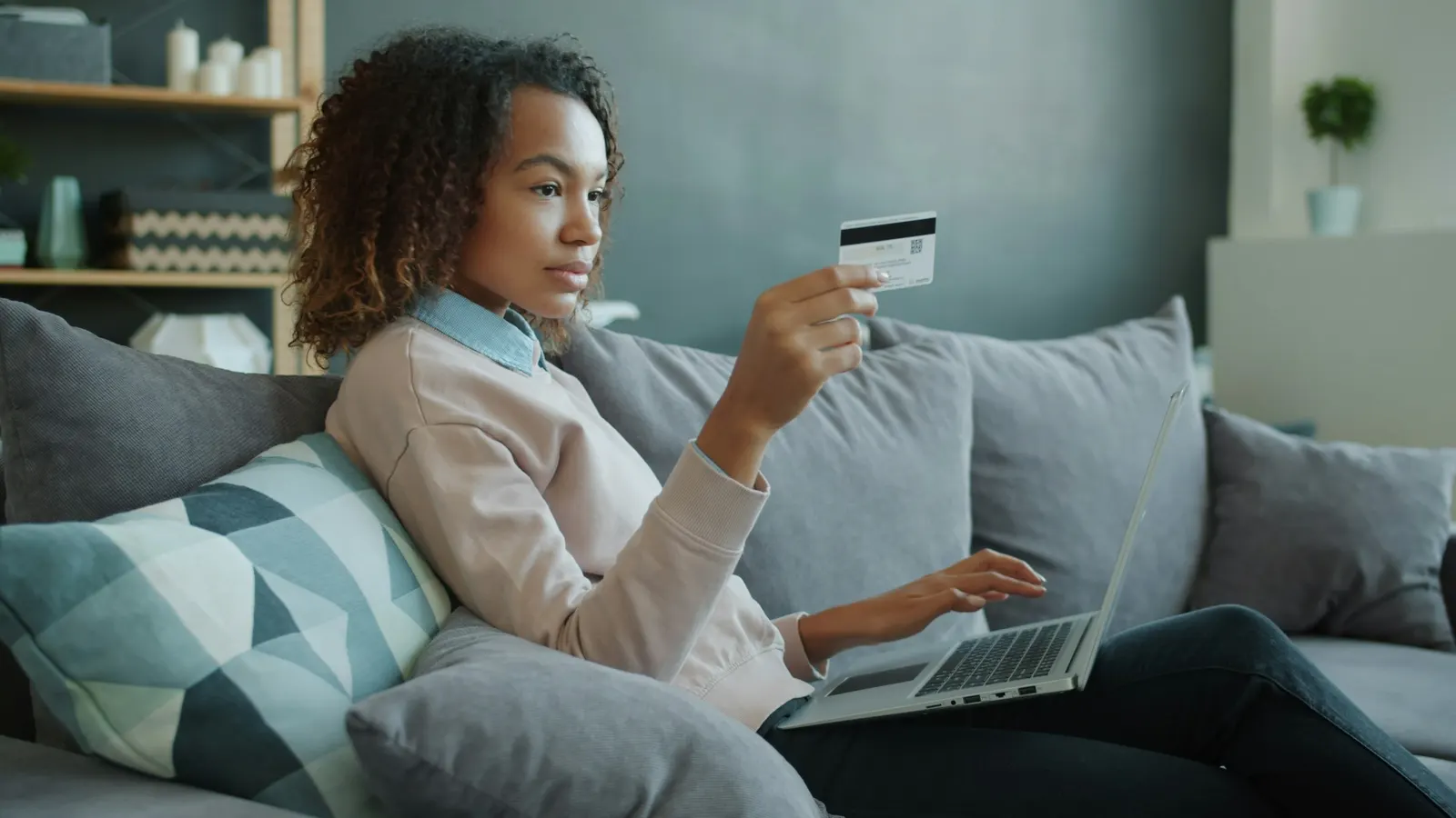 Woman on a sofa holding a payment card while typing on a laptop