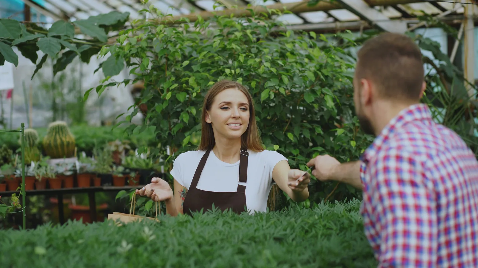 Customer handing a payment card to a smiling garden center worker in a greenhouse