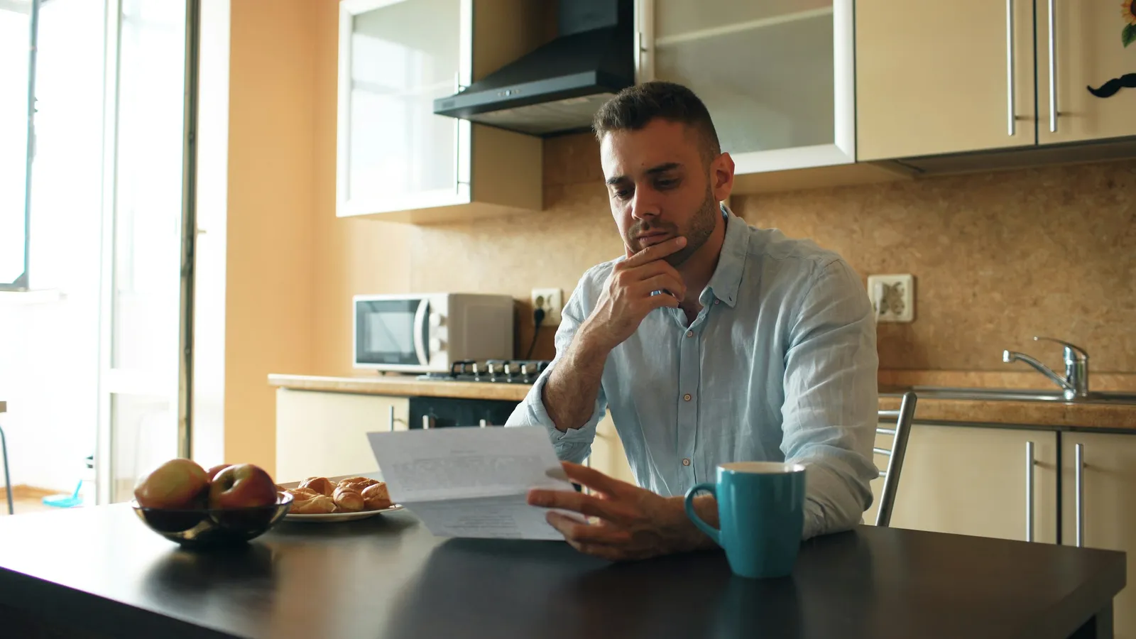 Man at a kitchen table reading a printed document, hand on his chin