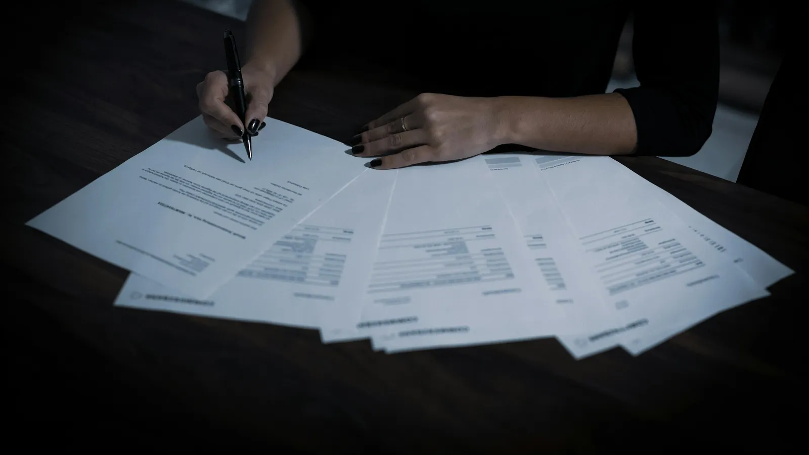 Woman signing printed documents spread across a dark table