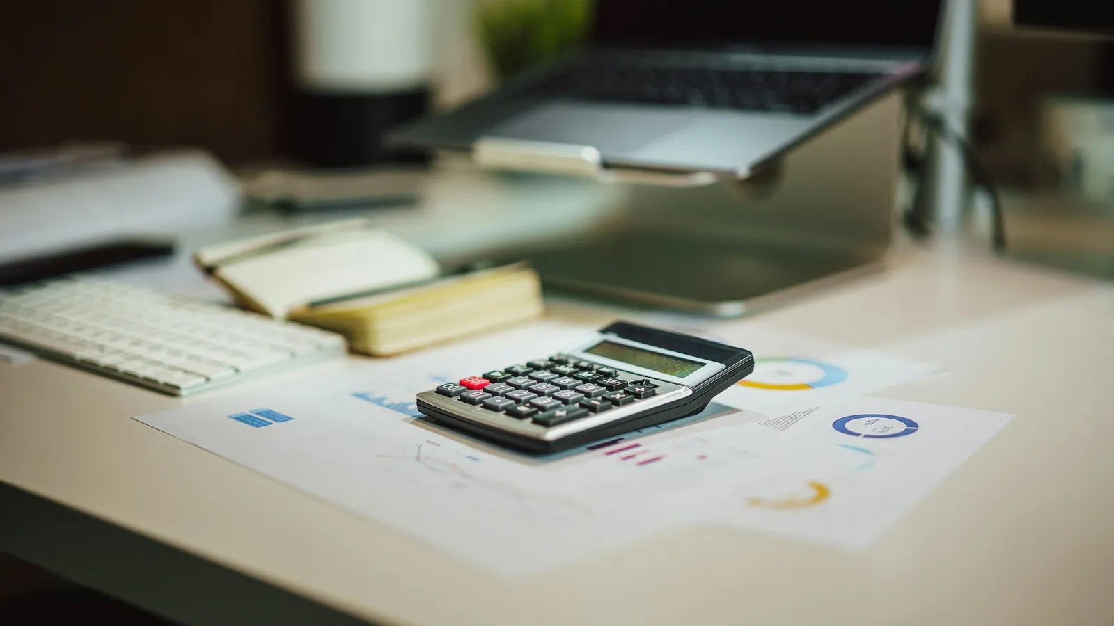 Calculator on printed charts on an office desk, with a laptop behind it
