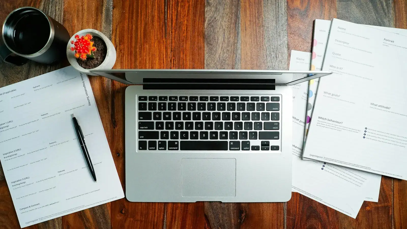 Open laptop on a wooden desk beside printed forms, a pen, a mug and a small cactus