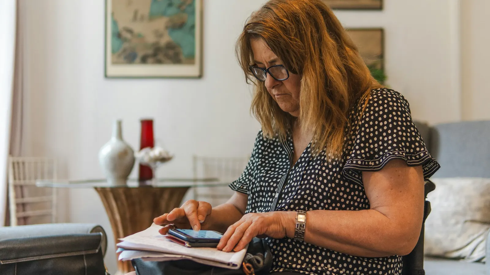 Woman in glasses tapping a smartphone resting on a stack of papers on her lap in a living room