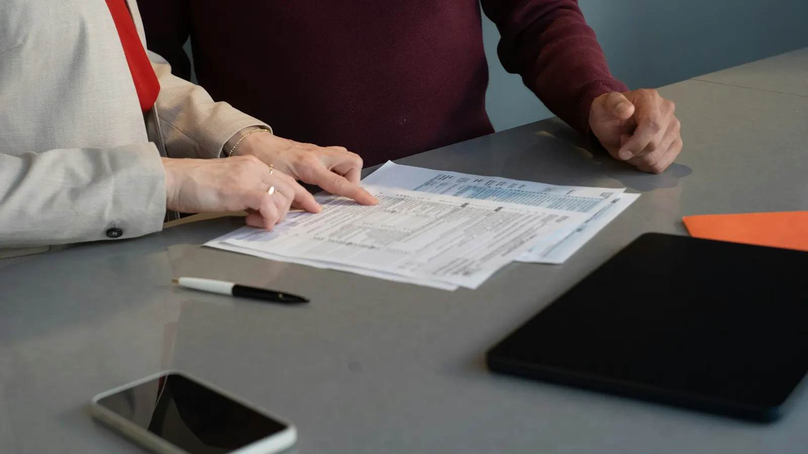 Two people at a table reviewing printed forms, with a pen, phone and tablet nearby
