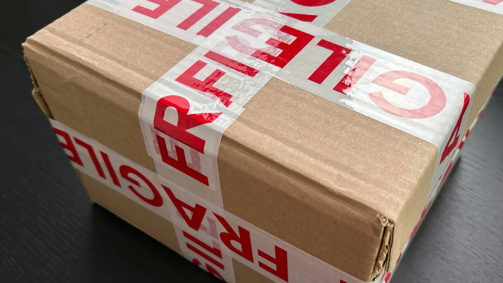 Cardboard shipping box sealed with red and white FRAGILE tape on a dark table